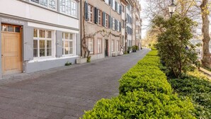 A paved street lined with buildings, a hedge, and a tree. - Marllon Suites – Casa Rheinblick Apartments – Bella Suite (Basel)