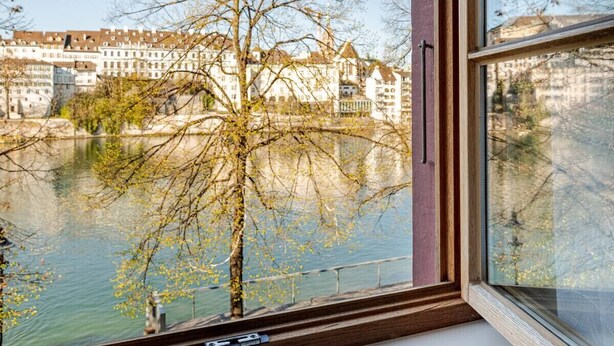 A window with a view of a river and buildings, a bare tree, and a wooden window frame. - Marllon Suites – Casa Rheinblick Apartments – Bella Suite (Basel)