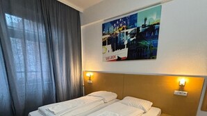 Un lit double avec des draps blancs soigneusement pliés, une tête de lit dotée de lampes de lecture intégrées et, au-dessus, un tableau représentant un paysage urbain.