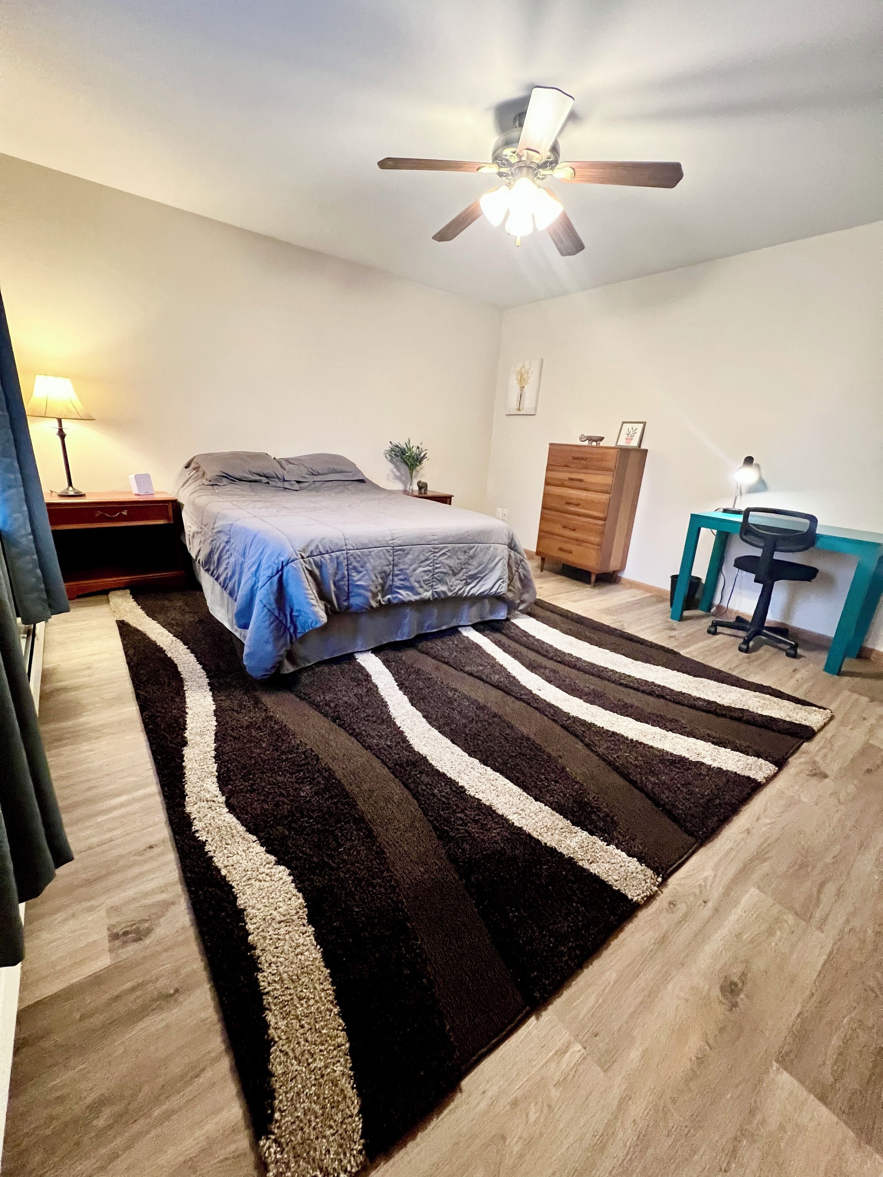 A bedroom with a bed, a desk, a chair, a dresser, a ceiling fan, and a striped rug.