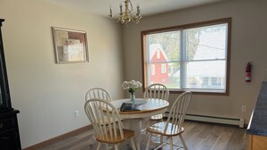 Een eetgedeelte met een ronde tafel, vier witte stoelen, een vaas met bloemen, een kroonluchter, een ingelijst schilderij en een raam met uitzicht op een rode woning.