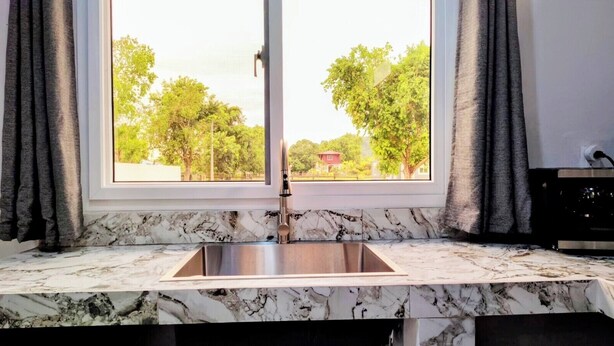Una cocina con encimera de mármol, pileta de acero inoxidable y ventana con cortinas.
