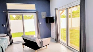 Un dormitorio moderno con cama, una silla, un televisor y amplias puertas correderas de vidrio que dan a un espacio exterior cubierto de césped.