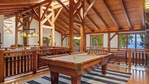 Uma mesa de bilhar em um salão com piso de madeira, teto alto e janelas amplas com vista para a água.