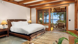 Quarto com cama de madeira, duas luminárias de cabeceira, vista do mar através de uma porta de vidro e um pequeno espaço de estar com mesa e cadeiras.