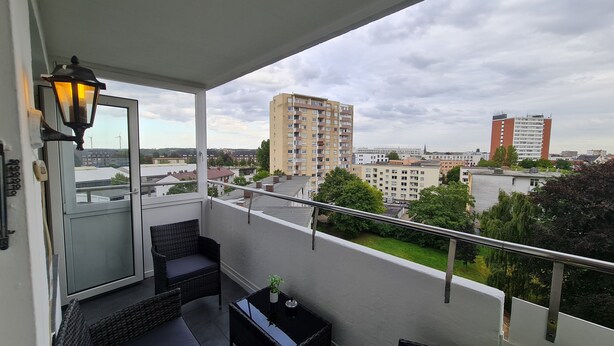 A balcony with a wicker chair, a small table, and a potted plant. It overlooks a cityscape with several buildings and a cloudy sky. - DEICHLIEBE 1 - fantastic view, own garage & contactless check-in (Bremerhaven)