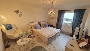 A bedroom with a bed, bedside tables, a chair, a small table with a vase, and a view of the outside through a window. - DEICHLIEBE 1 - fantastic view, own garage & contactless check-in (Bremerhaven)