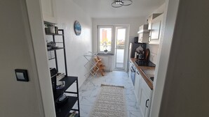 A narrow hallway leading to a kitchen with white cabinets, a black countertop, and a window with a view of the outside. - DEICHLIEBE 1 - fantastic view, own garage & contactless check-in (Bremerhaven)