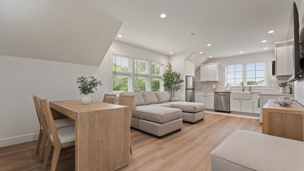Eine moderne Küche mit weißen Schränken, ein Essbereich mit Holztisch und -stühlen sowie ein Wohnbereich mit Sofa und Hocker.