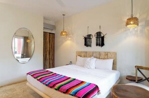 Une chambre d’hôtel avec un grand lit, un miroir rond, deux lampes fixées au mur et une chaise en bois.