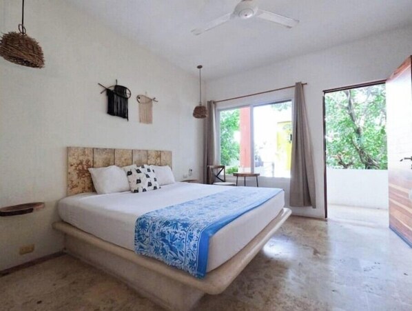 Une chambre avec un lit, un ventilateur de plafond, une fenêtre et une vue sur la verdure environnante.