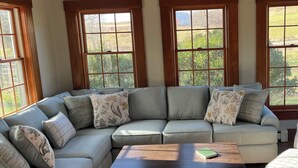 A L-shaped sectional sofa with floral cushions, a wooden coffee table, and large windows with a view of greenery. - Large Farmhouse with expansive views, firepit 15 min north of Keene (Walpole)