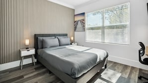 A modern bedroom with a large bed, a nightstand, a wall-mounted lamp, a window with blinds, and a ceiling fan. - The Heights Collective (Tampa)