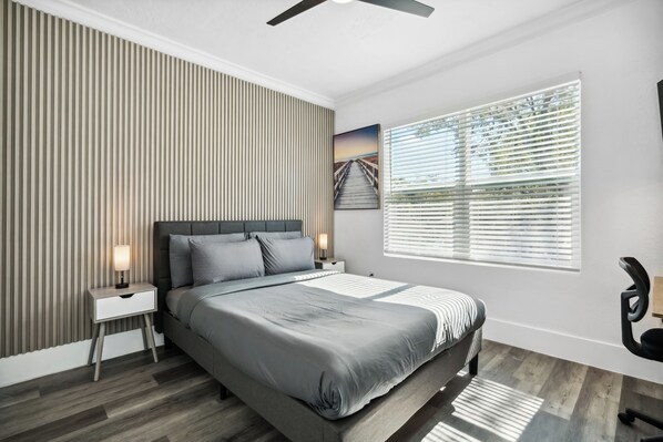 A modern bedroom with a large bed, a nightstand, a wall-mounted lamp, a window with blinds, and a ceiling fan.