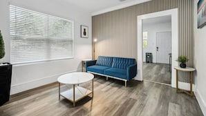 A modern living room with a blue sofa, a round coffee table, and a large window with blinds. - The Heights Collective (Tampa)