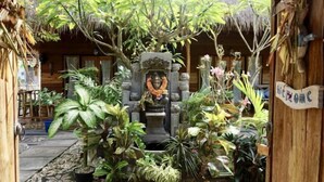 Una estructura de madera tradicional con techo de paja, rodeada de exuberante vegetación tropical y una estatua de piedra en el centro.