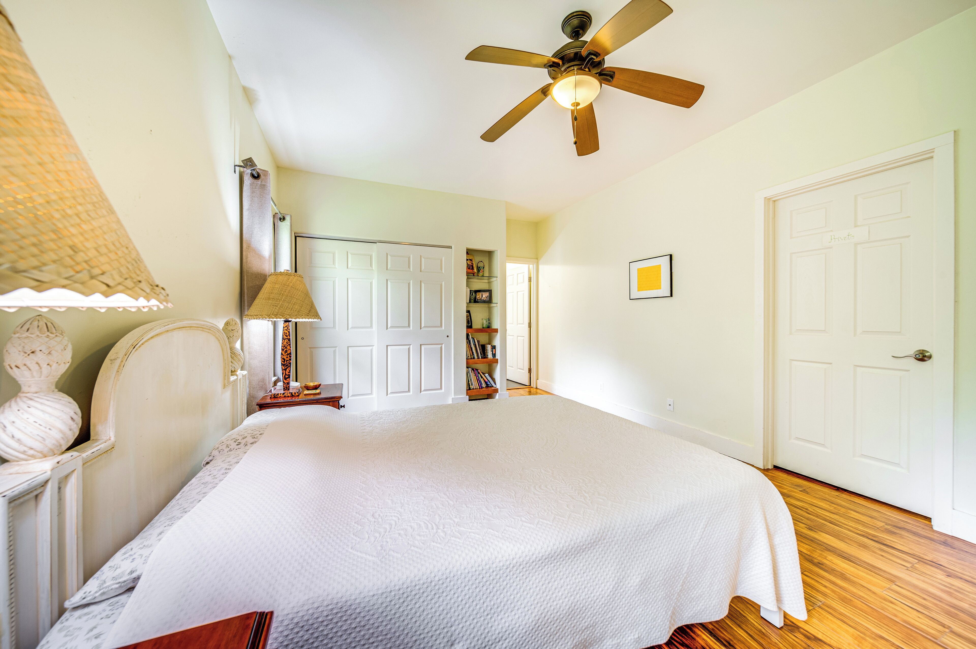 Une chambre à coucher avec un lit, un ventilateur de plafond, une lampe, une bibliothèque et une porte.