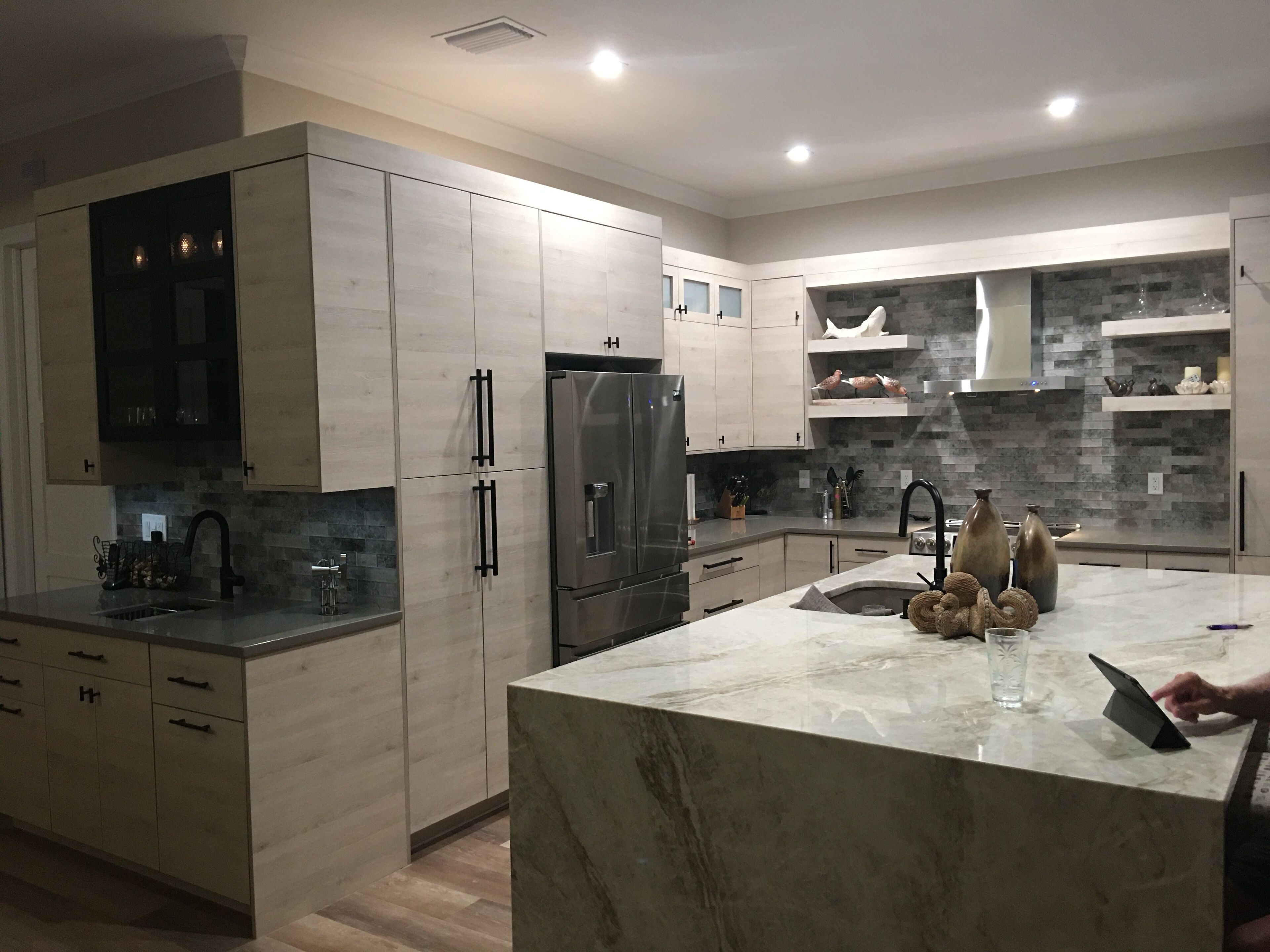 A modern kitchen with light wood cabinetry, a stainless steel refrigerator, and a marble island.