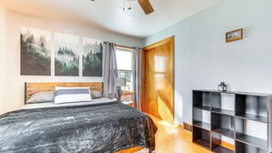 Een slaapkamer met een bed, een houten hoofdeinde, een plafondventilator, een raam met gordijnen, een boekenkast en een ingelijst schilderij aan de muur.