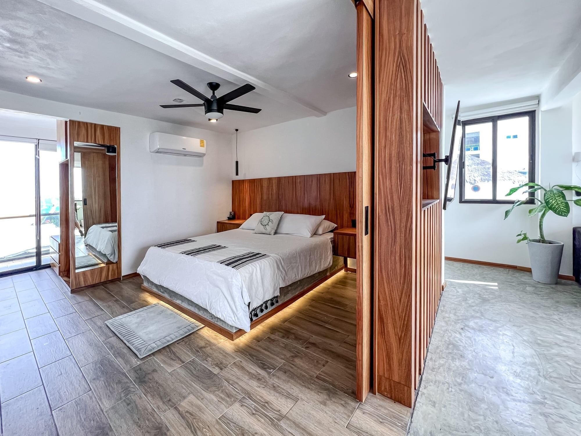 Ein modernes Schlafzimmer mit einem Holz-Kopfteil, einem Bett mit weißer Bettwäsche, einem Deckenventilator und einem großen Fenster.