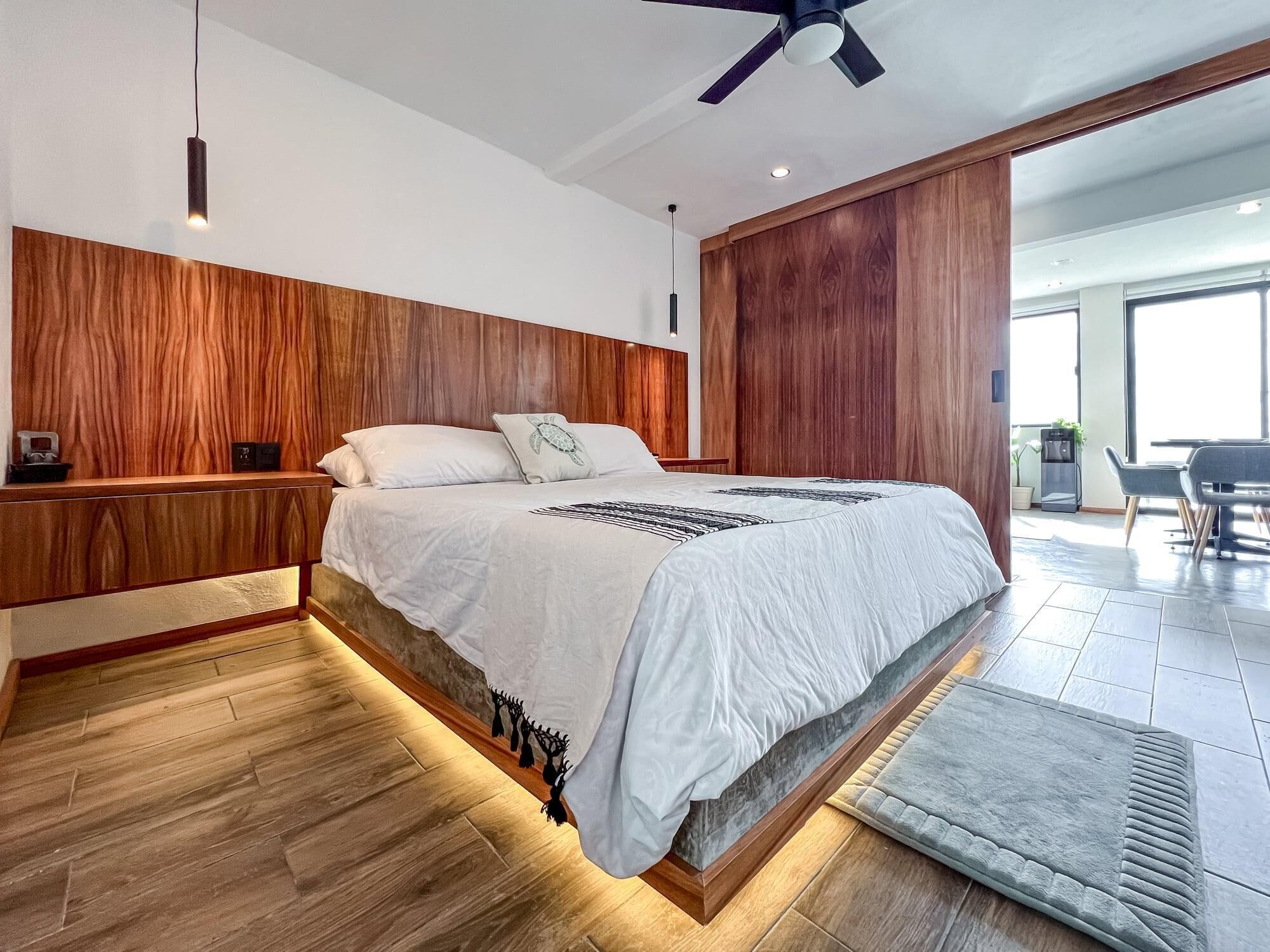 A modern bedroom with a wooden headboard, a large bed with white bedding, a ceiling fan, and a view of the living area through a glass door.