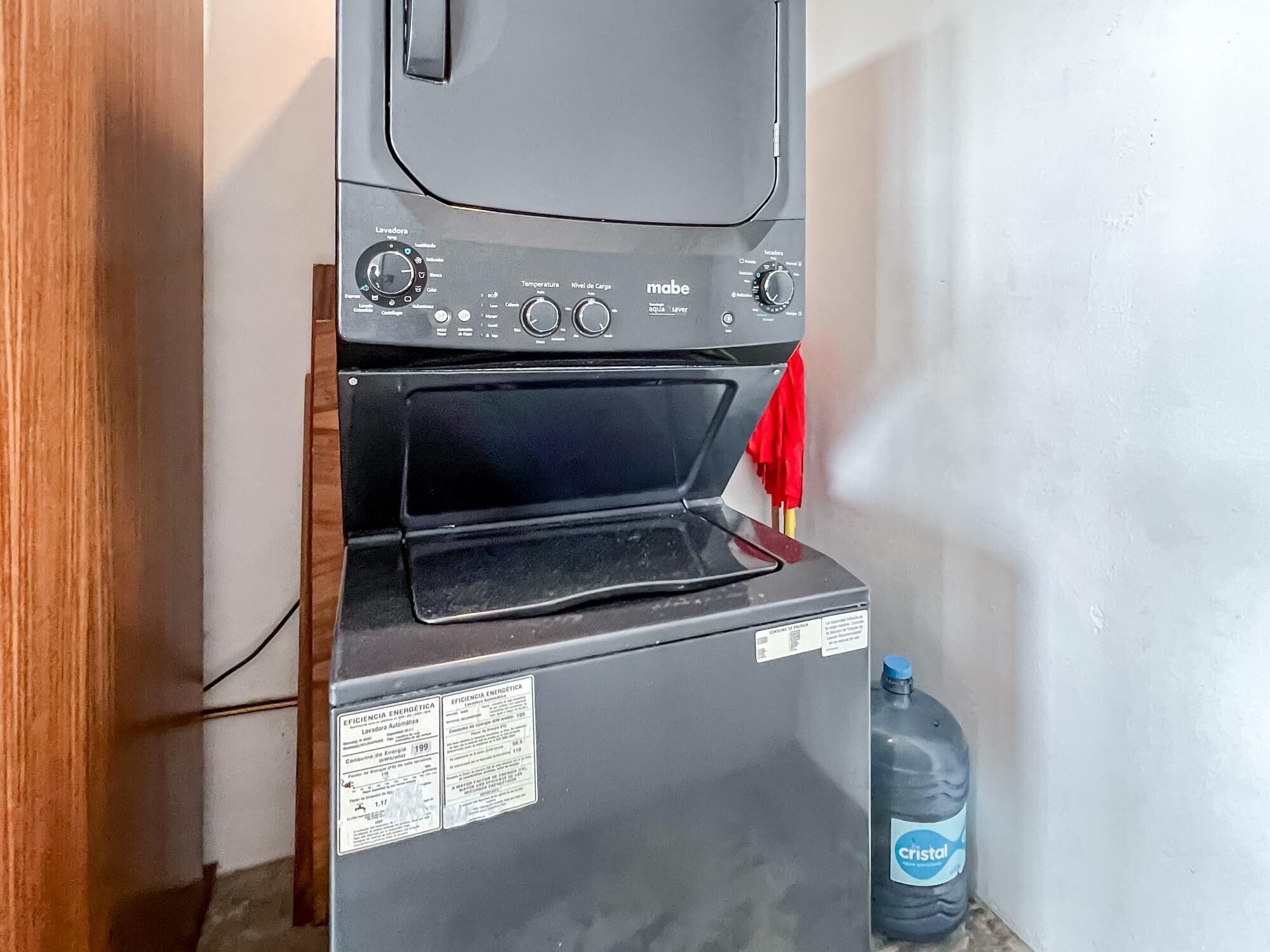 Een compacte, zwarte wasmachine met een voorlaadklep, een bedieningspaneel met verschillende standen en een waterfles met de tekst 'Cristal' ernaast.