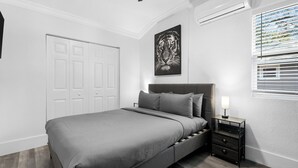 A bedroom with a bed, a nightstand, a wall-mounted lamp, a door, and a window with blinds. - The Heights Collective (Tampa)