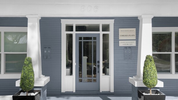 A building entrance with a gray facade, a white door, and two potted topiary trees. - The Heights Collective (Tampa)