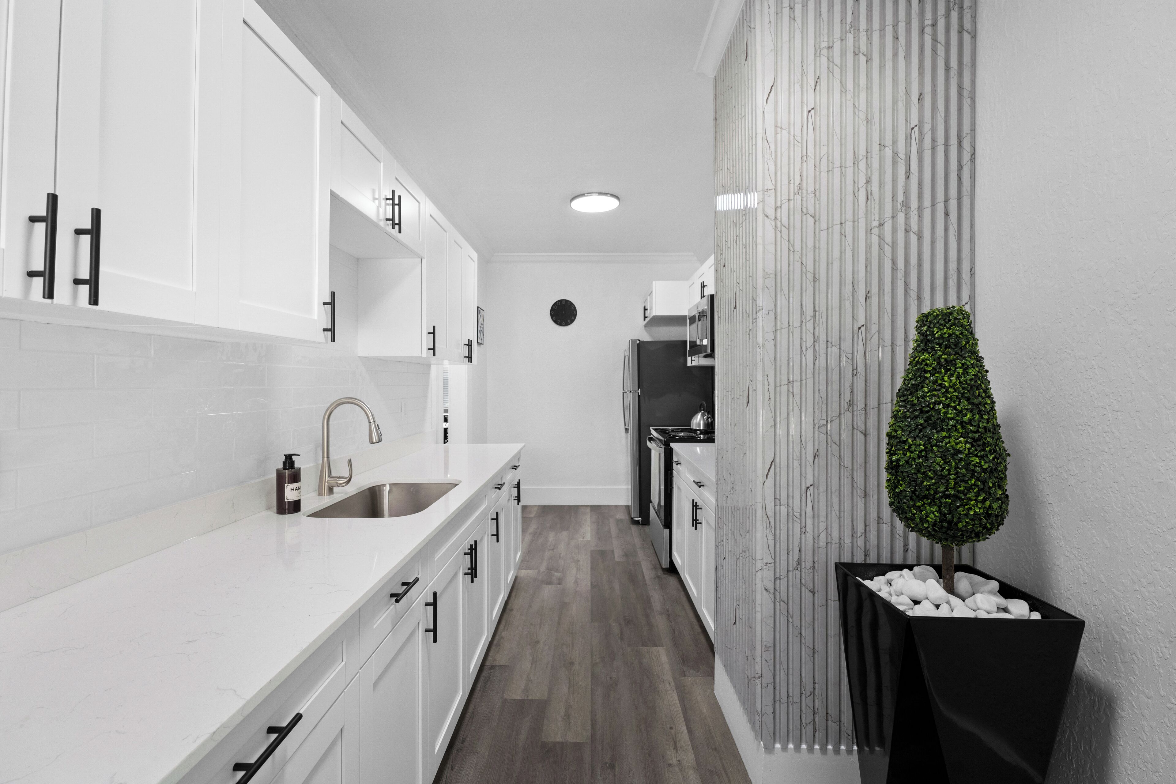 A narrow kitchen with white cabinets, a stainless steel sink, and a potted plant.