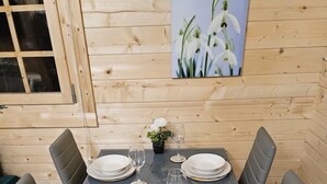 Una mesa de comedor dispuesta con platos, vasos y cubiertos, rodeada de sillas, en una habitación con paredes de madera y un cuadro de flores.