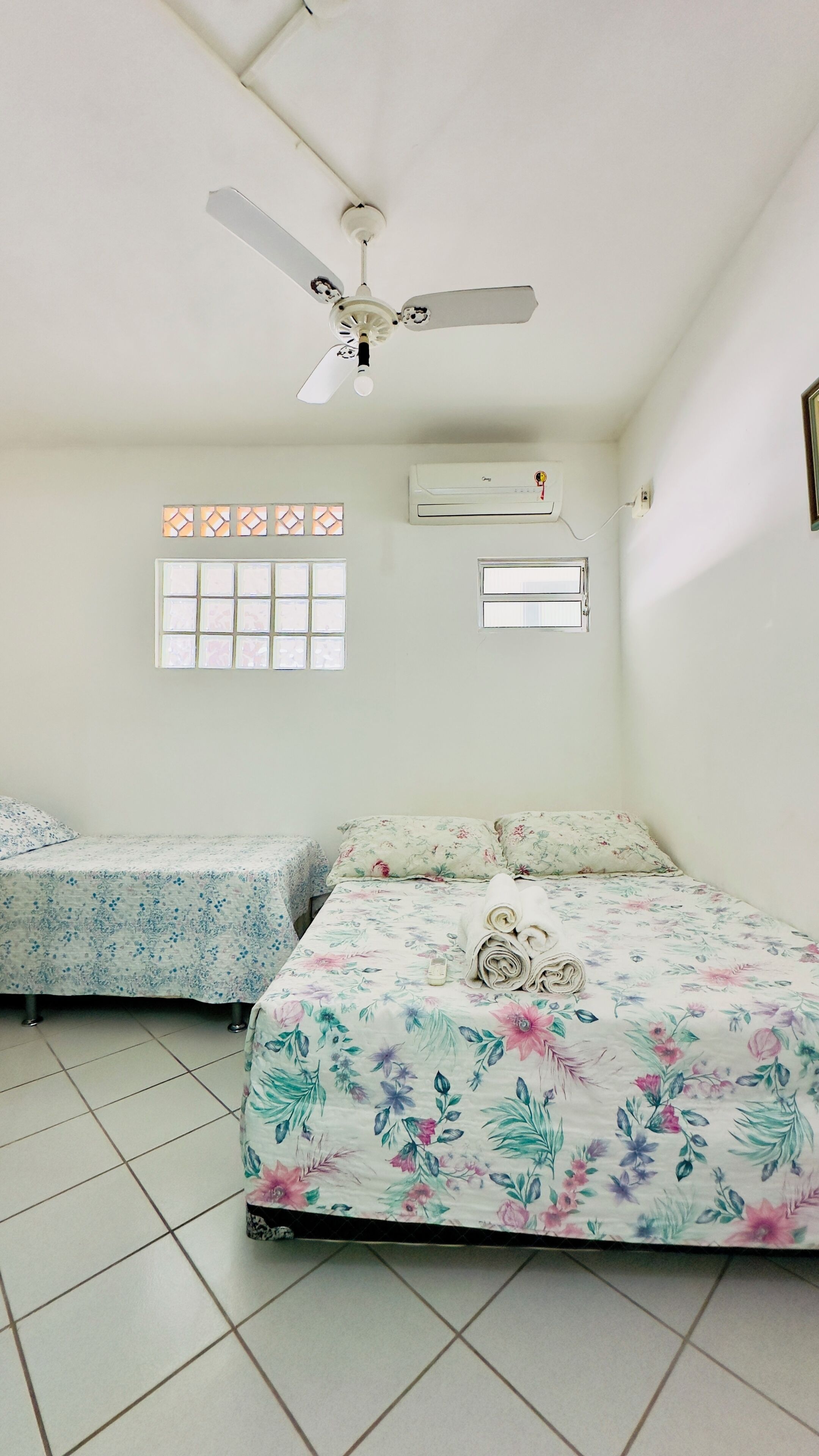 A room with two beds, a ceiling fan, an air conditioning unit, and a window with a decorative grid.