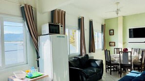Un saló amb un sofà de pell negra, un refrigerador blanc, una taula de dinar amb cadires i un televisor fixat a la paret.