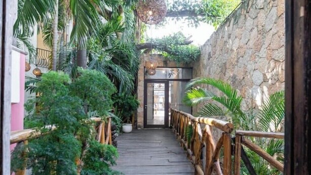 Ein Holzweg führt zu einem Gebäudeeingang, umgeben von üppiger Bepflanzung und Steinmauern.