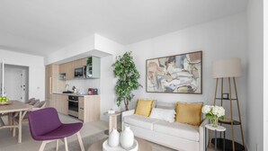 A modern living room with a white sofa, a purple chair, and a coffee table. There is a kitchen in the background with a microwave and cabinets.