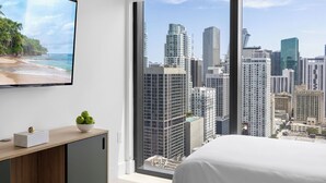 A bedroom with a large window offering a cityscape view, a bed, a bedside table with a bowl of apples, and a flat-screen TV on the wall.