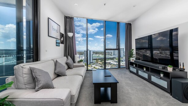 A modern living room with a sofa, a coffee table, a TV, and a view of a cityscape through large windows.