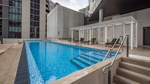 A modern indoor swimming pool with clear blue water, surrounded by lounge chairs and a covered seating area with patterned walls.