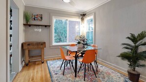 Ein Essbereich mit einem Glastisch, orangefarbenen Stühlen, einem Holzsideboard, einem Deckenventilator, einem Fenster mit Blick auf Bäume und einer Topfpflanze.