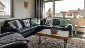 Uma sala de estar moderna com um sofá em formato de L, uma mesa de centro e vista para o exterior através de grandes janelas.