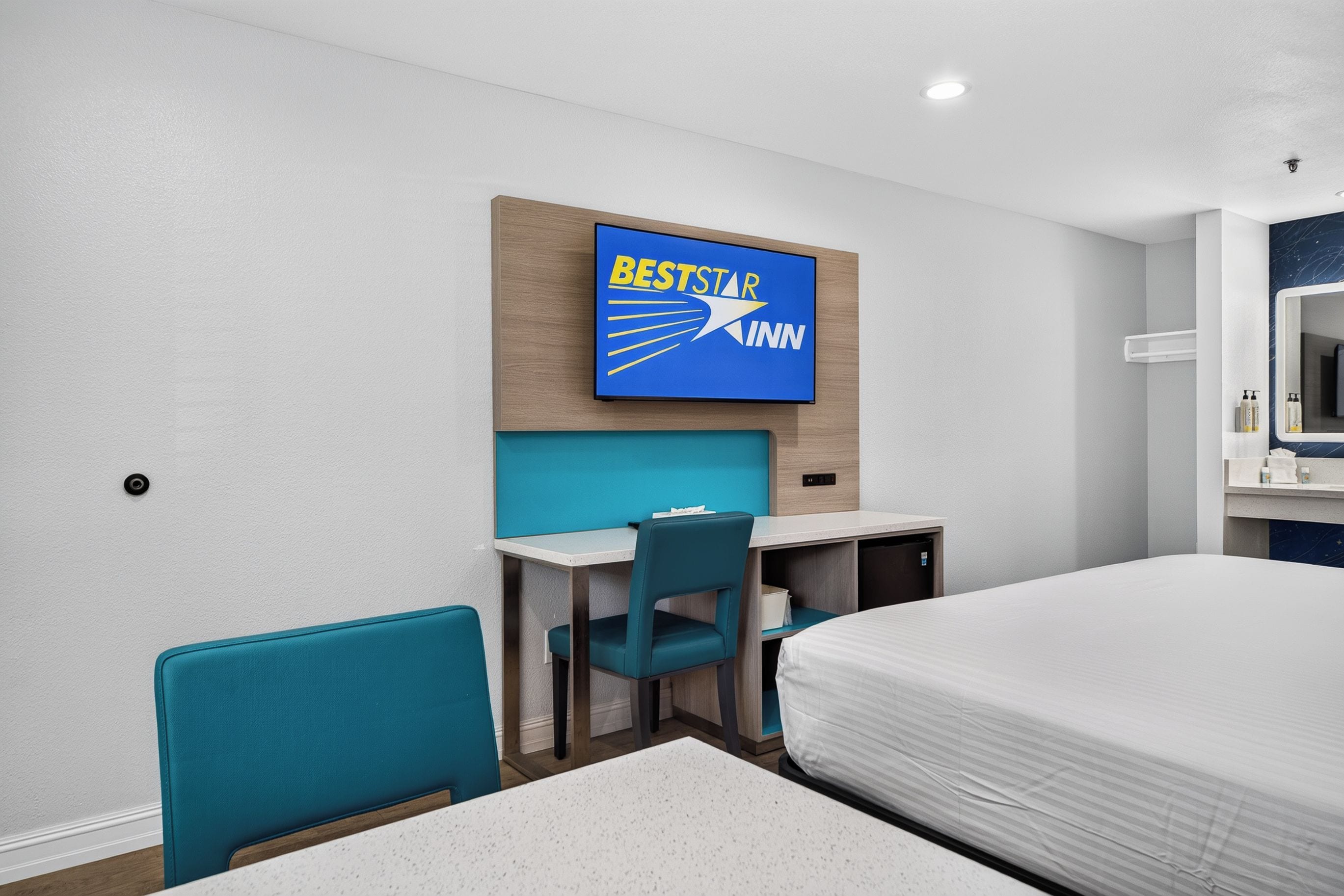 Habitación de hotel con cama, escritorio, dos sillas, televisor montado en la pared y baño con espejo y toallero.
