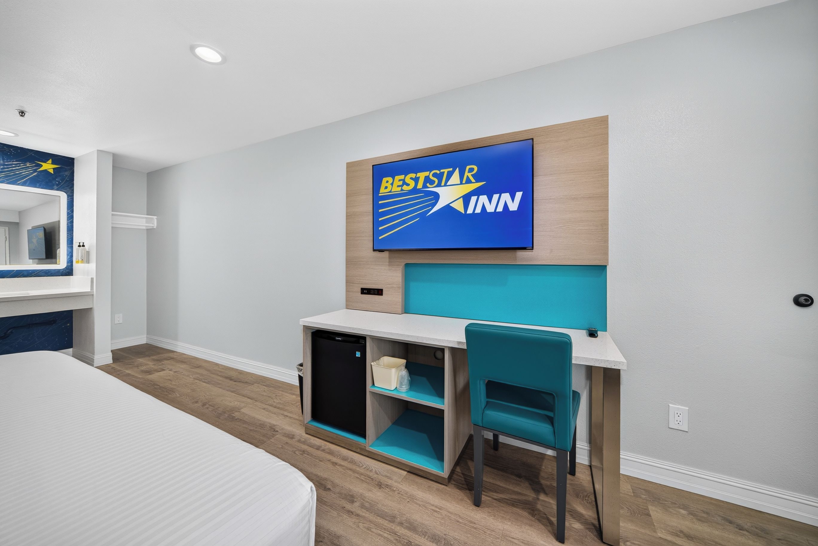 A hotel room with a bed, a desk with a chair, a television mounted on the wall, and a small kitchenette area.