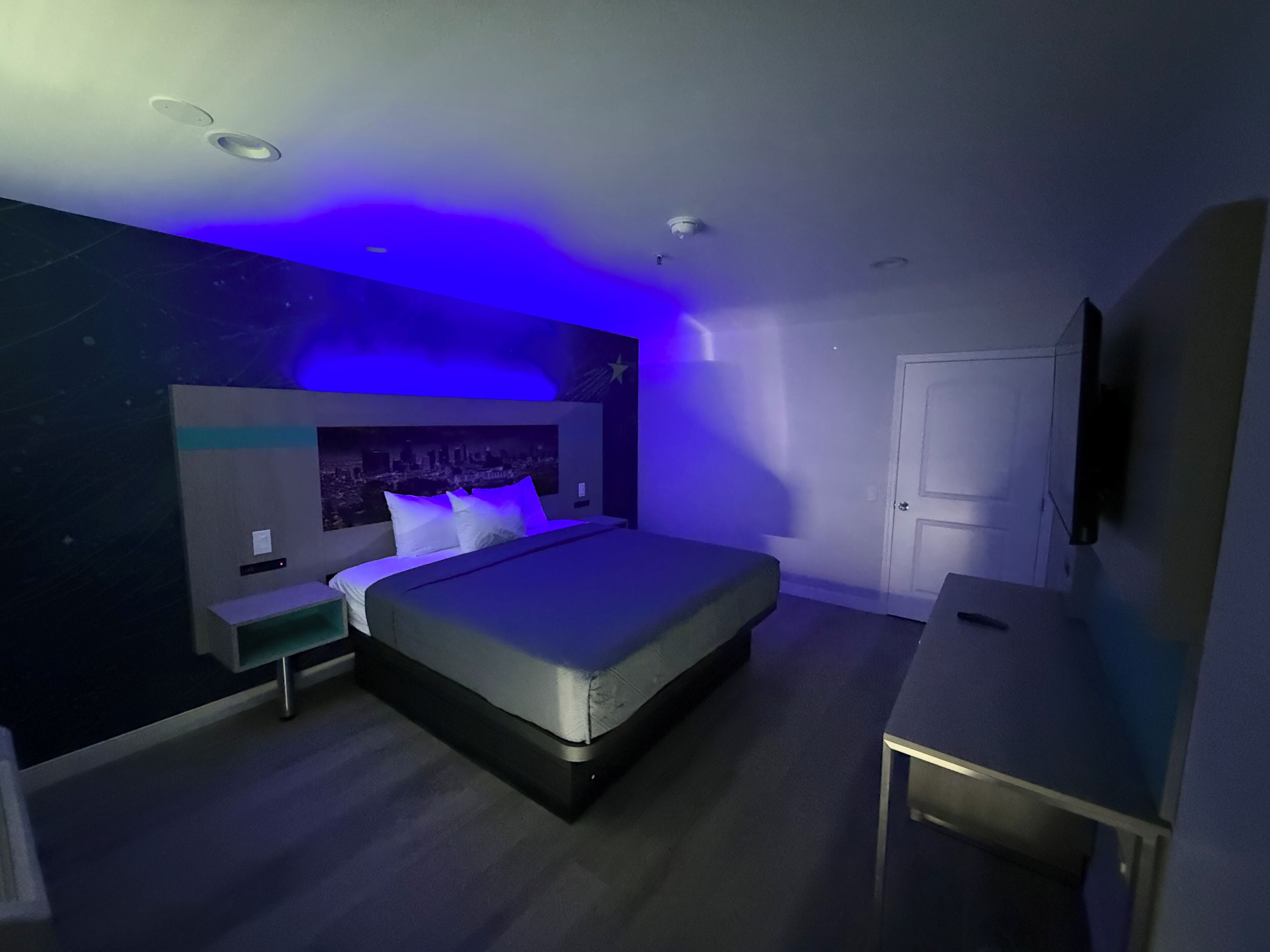 Ein modernes Schlafzimmer mit Stadtbild-Wandgemälde, einem Bett mit weißen Bettwäsche, einem Nachttisch und einem Schreibtisch mit Stuhl.