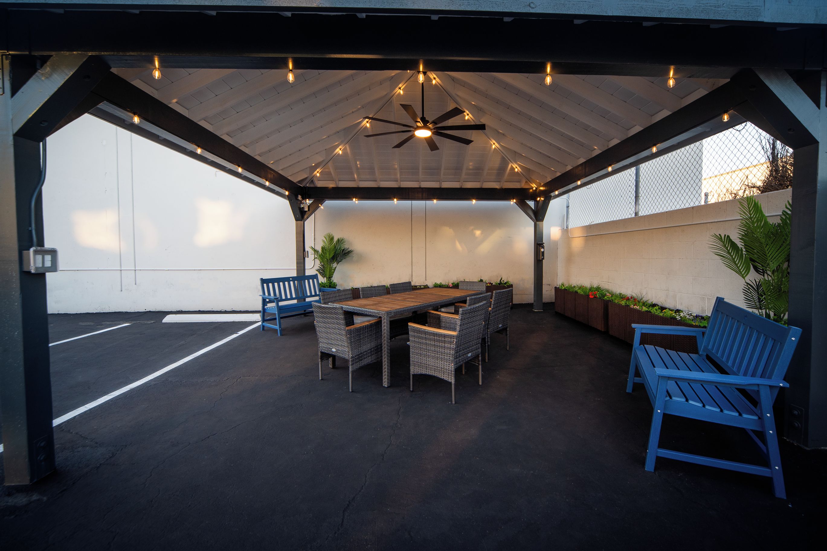 An outdoor patio with a ceiling fan, string lights, a wooden table, wicker chairs, and a blue bench.