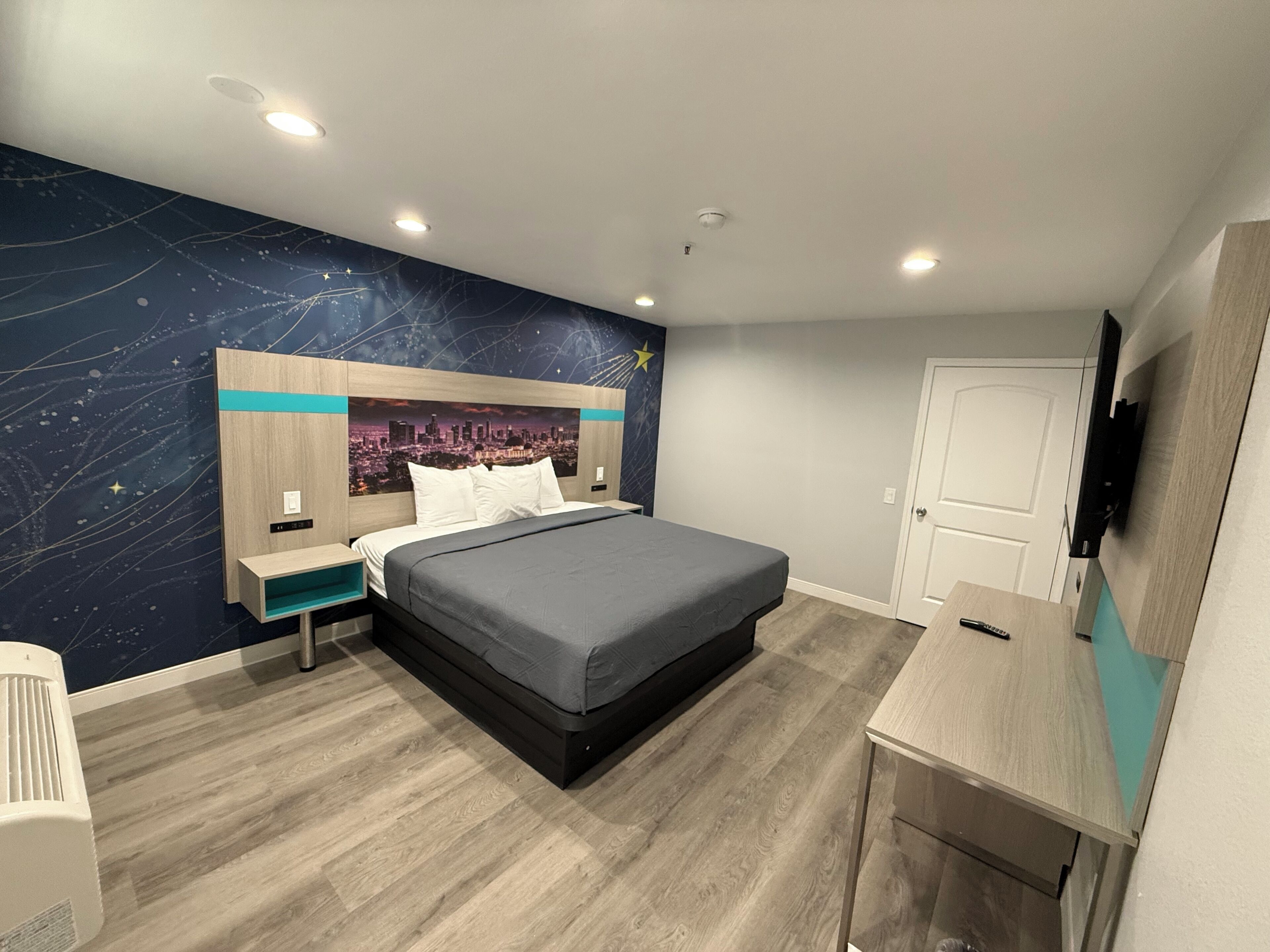 A modern hotel room with a bed, a desk, a TV, and a cityscape mural on the wall.