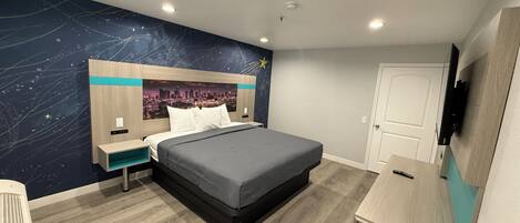 A modern hotel room with a bed, a desk, a TV, and a cityscape mural on the wall.