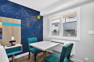Una habitación con un mural de estrellas en la pared, una mesa con sillas, una ventana con persianas y un pequeño escritorio con un teléfono.