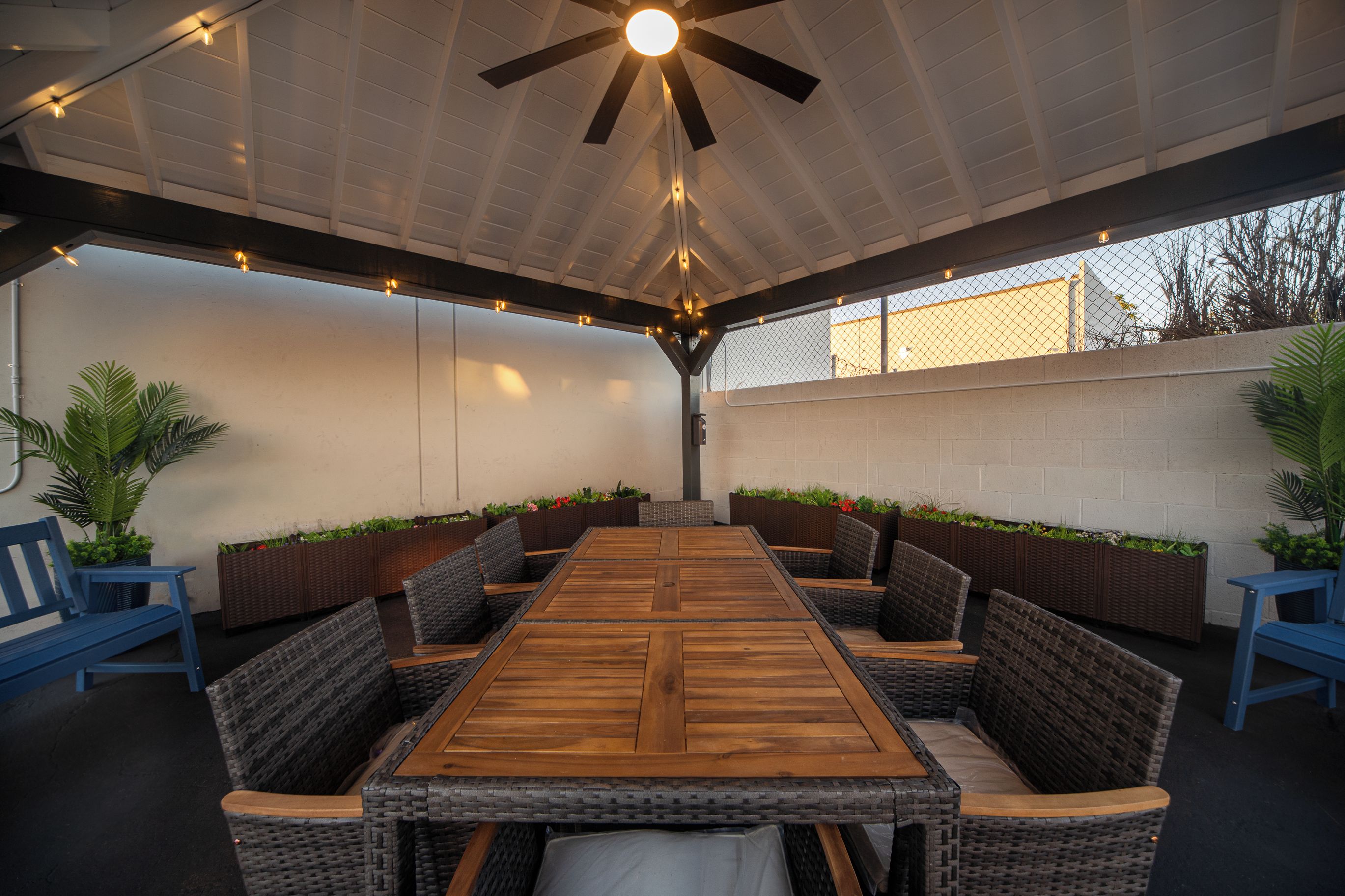 An outdoor patio with a wooden table surrounded by wicker chairs, a ceiling fan, and string lights.