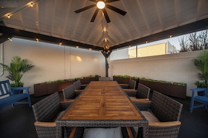 Un patio exterior con una mesa de madera rodeada de sillas de mimbre, un ventilador de techo y luces colgantes.