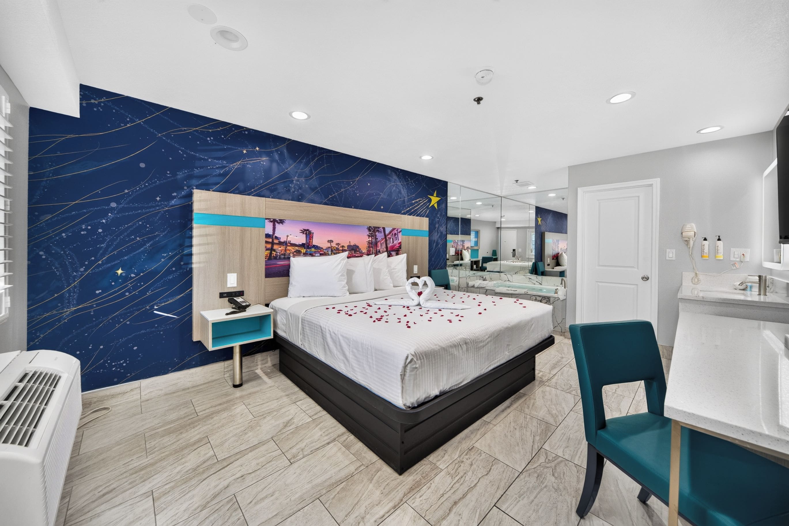 A modern hotel room with a large bed, a nightstand, a desk, a chair, a bathroom with a mirror, and a wall with a starry night design.