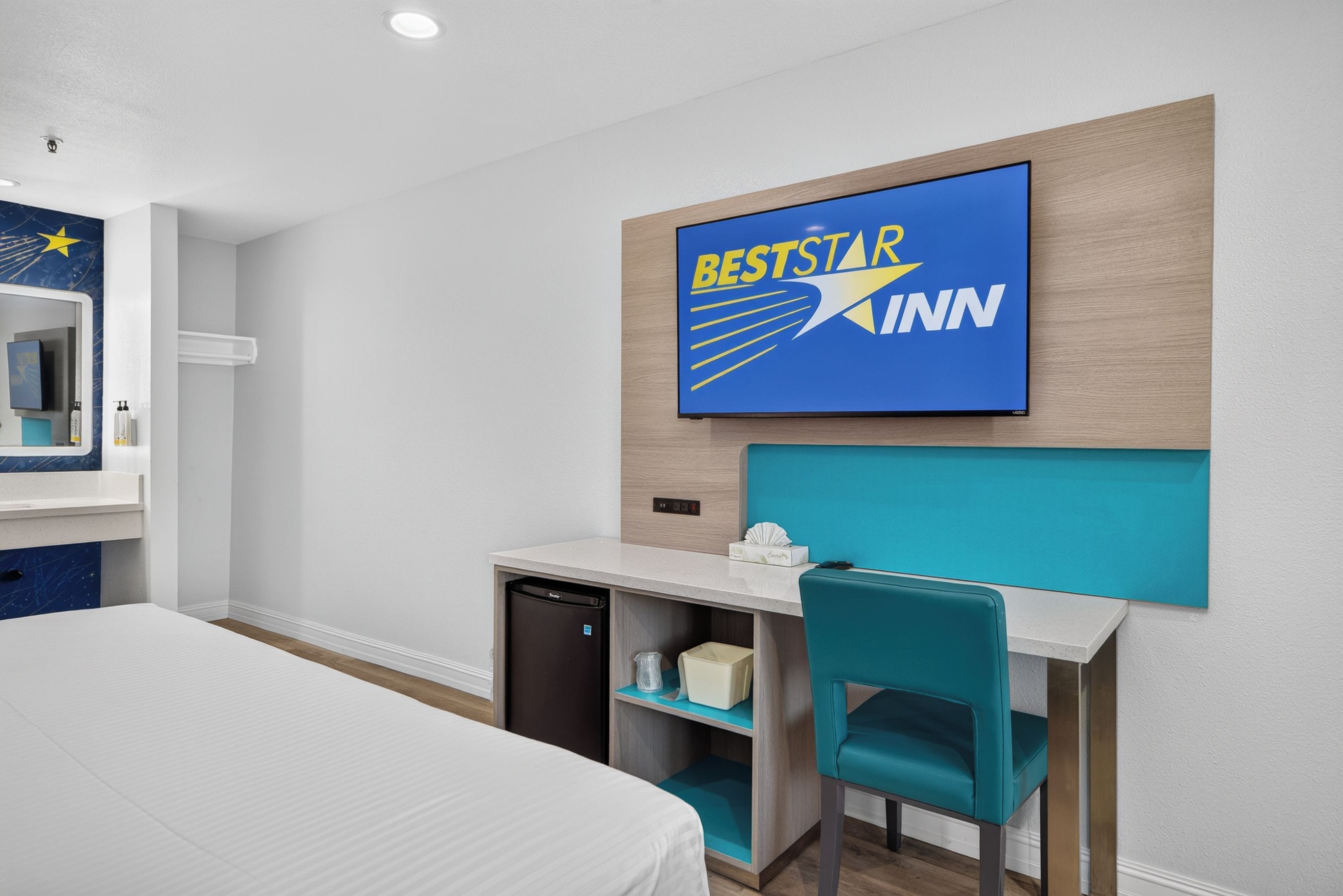 A hotel room with a bed, a desk with a chair, a small refrigerator, a wall-mounted TV displaying 'BESTSTAR KINN', and a mirror with toiletries.